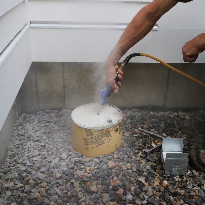 Hand holding compressor hose blowing dust out of the hole drilled for post-base anchors.