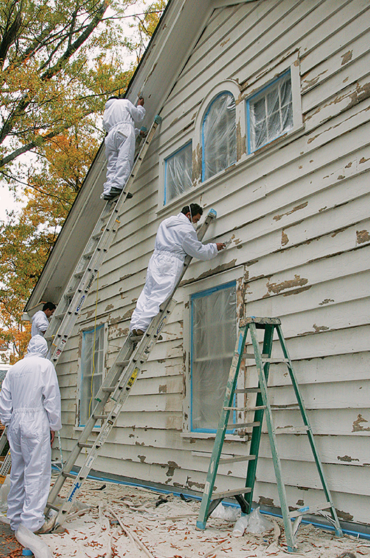 Photo of three men addressing peeling paint on the side of a house.